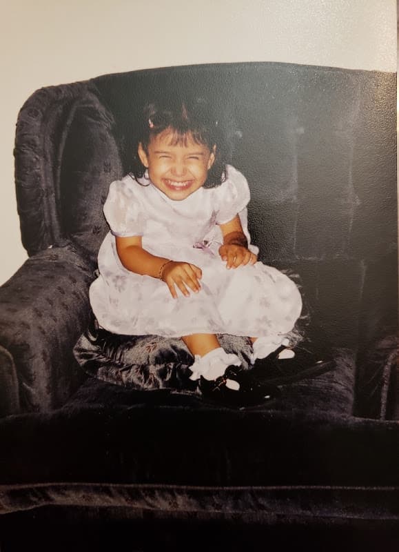 Jenn as a toddler, smiling in a white dress